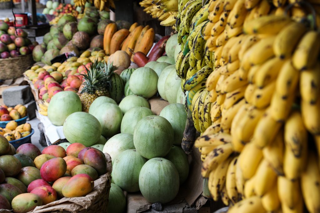 Several racimes of bananas hang in the foreground at a roadside food stall in Guatemala. A basket of mangos and stacks of melons are in focus behind the bananas.