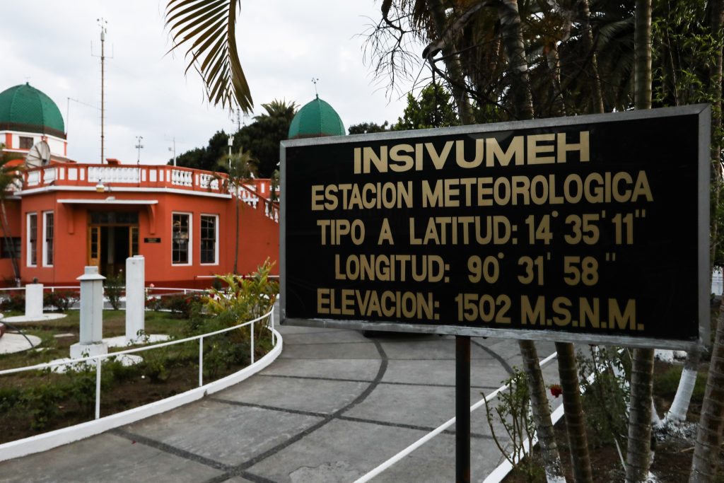 This photo captures the exterior of the Guatemala City office of Guatemala's meteorological agency. The building is single story with an orange exterior, white railings and two green domes perched on the roof with a garden in front. A sign in the foreground of the photo identifies the building as a meteorological station as well as the latitude, longitude, and elevation (in Spanish).
