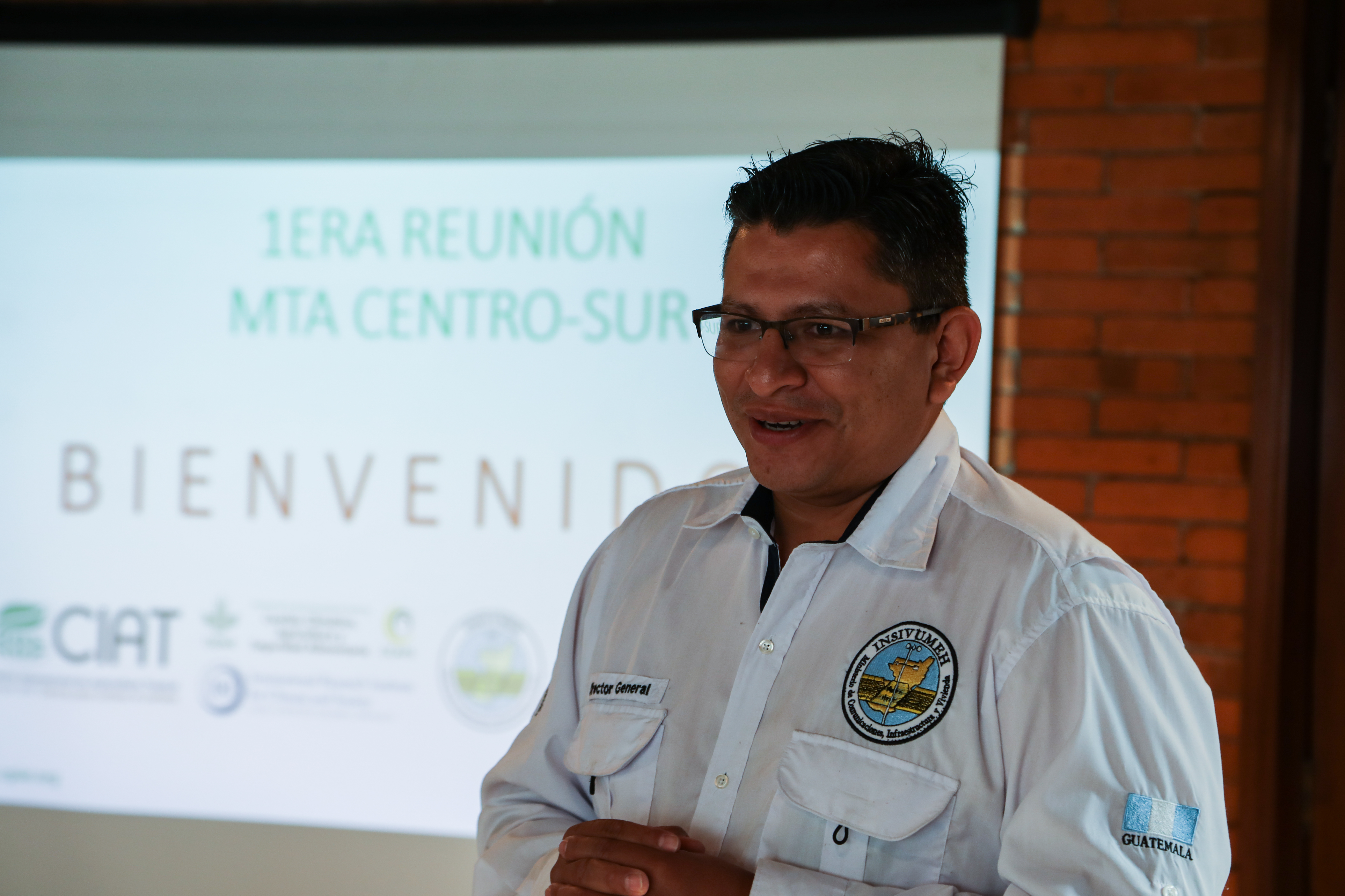 Juan Pablo Oliva Hernández, a Guatemalan man wearing glases and a button-up shirt with the INSIVUMEH logo, stands in front of a projector screen.