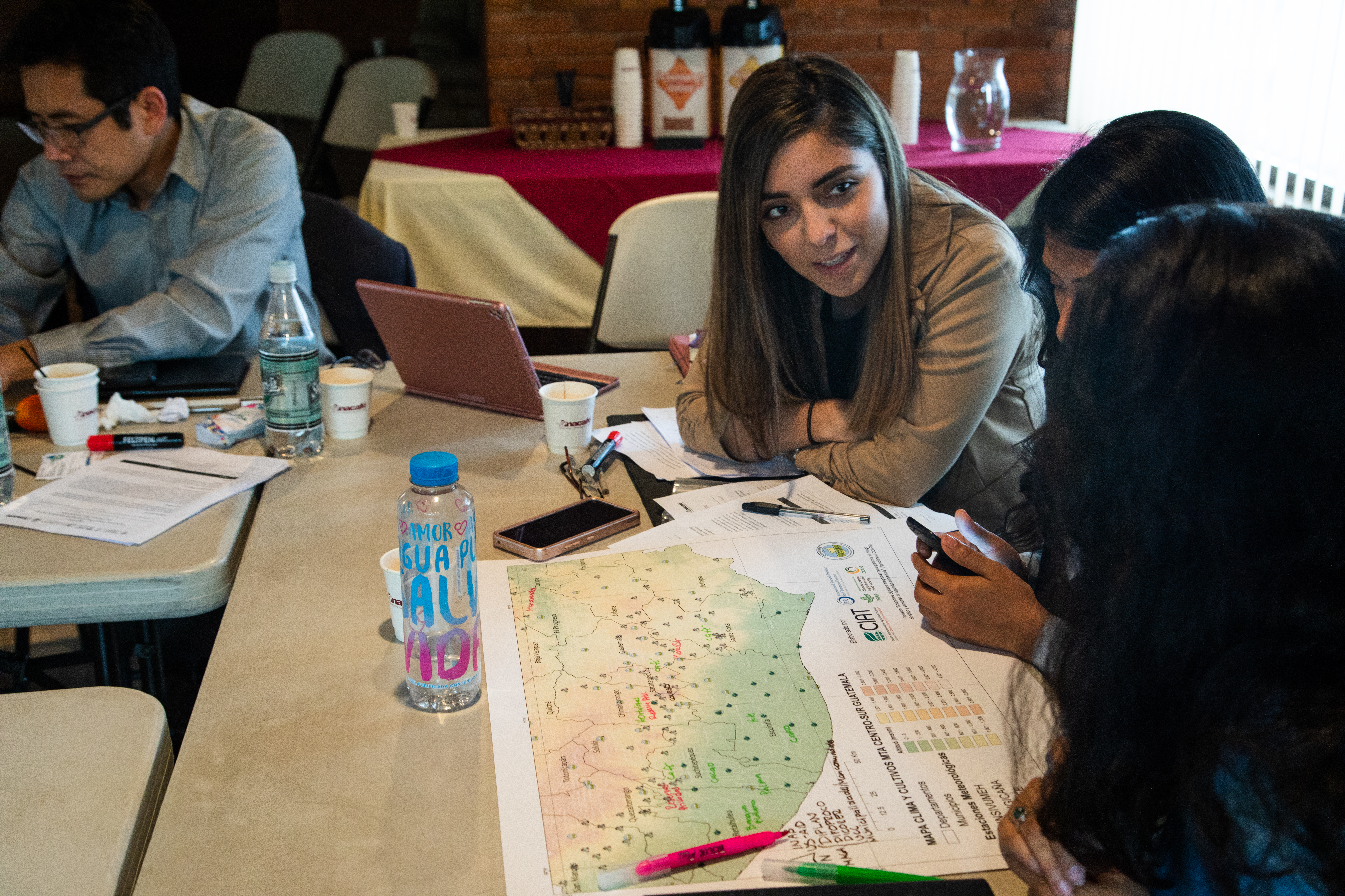 Three Guatemalan women lean over a map of southern Guatemala placed on a folding table in a meeting room. Only one woman's face faces the camera. Their map has highlighter and pen markings on it and they are in deep discussion.