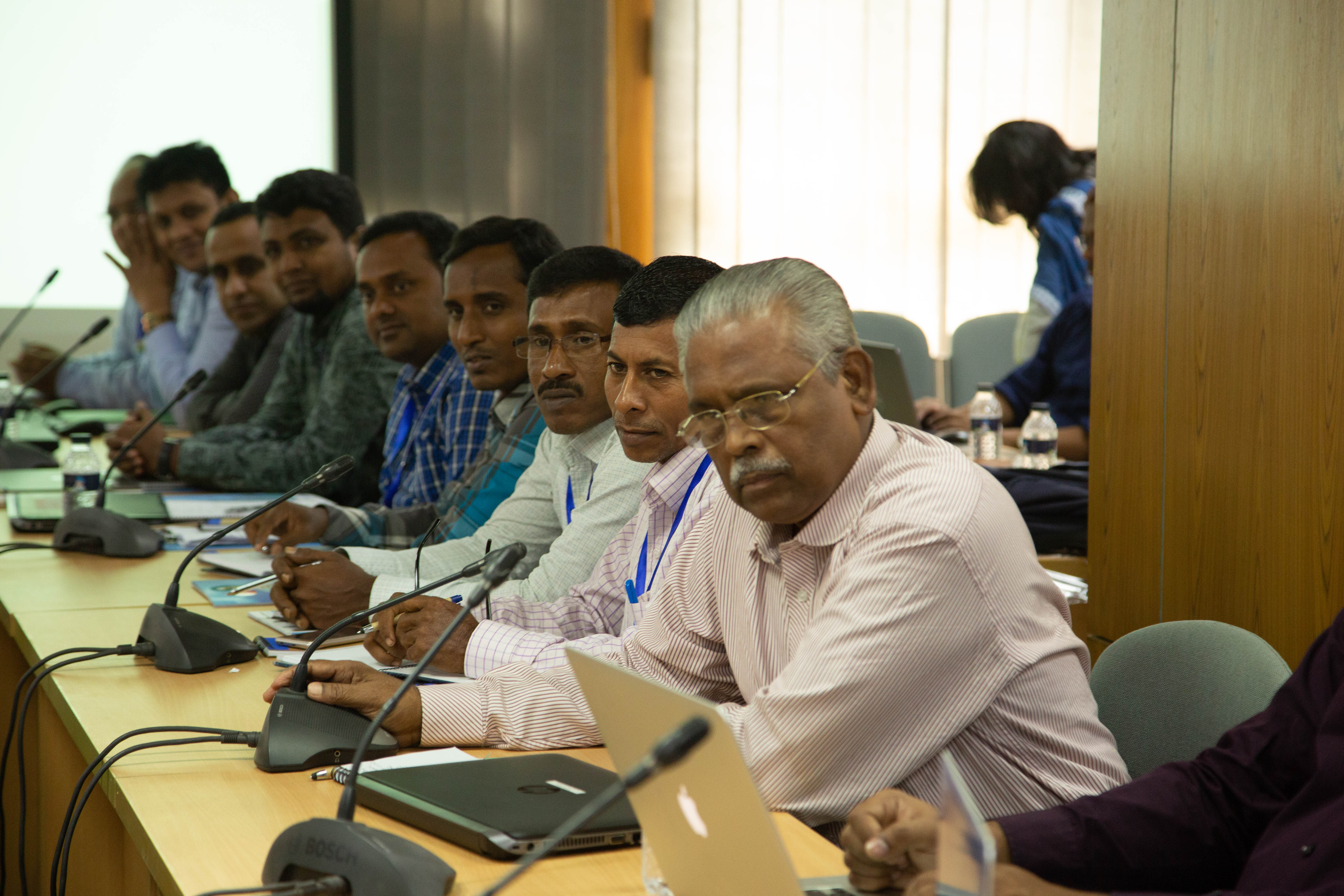 A row of Bangladeshi men of varying ages sit at a conference table with microphones and notepads in front of them. They are participants listening to a presentation during a training held at the Bangladesh Meteorological Department in Dhaka in October of 2019. Photographer Jacquelyn Turner, ACToday/IRI.