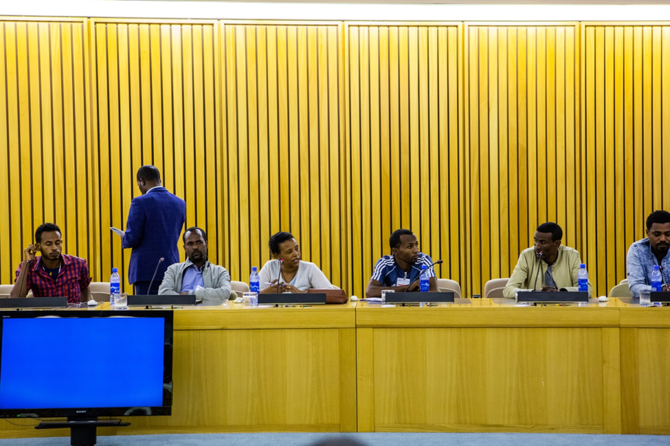 Eneye Assefa, third seated from the left, speaks during the closing ceremony of the training. Wood paneling stretches up the wall behind the panel of six Ethiopian participants sitting at the panel-like table. Each has a plastic water bottle and microphone in front of them. Photographer Elisabeth Gawthrop, ACToday/IRI.