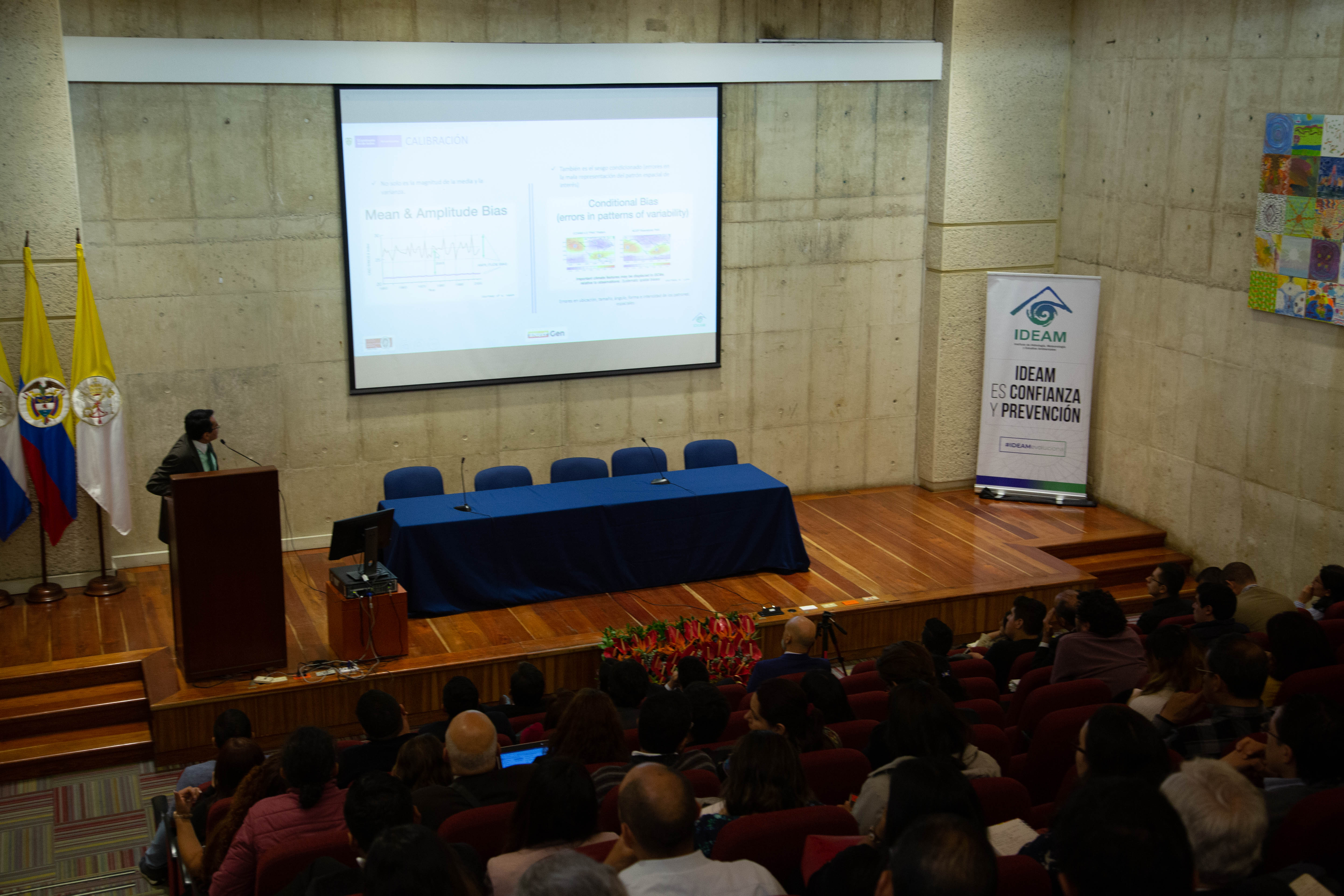 José Franklyn Ruiz, the Head of Weather and Climate Modeling Group for the National Meteorological Service of Colombia, stands behind a podium at the front of a lecture hall with concrete walls at the NextGen launch event on August 13th, 2019. On the wooden stage in front of the screen is a Colombian flag and an empty panel table covered with a blue table cloth. The audience seating is full. Photographer Jacquelyn Turner, ACToday/IRI.